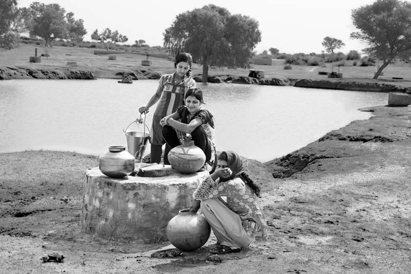 Portrayal of prosperity in Binjharwali village where young girls chat on the mobile phone while happily collecting drinking water from a shallow well (kuin) situated within the village catchment (paitan) - village Binjharwali, Bikaner district, Rajasthan